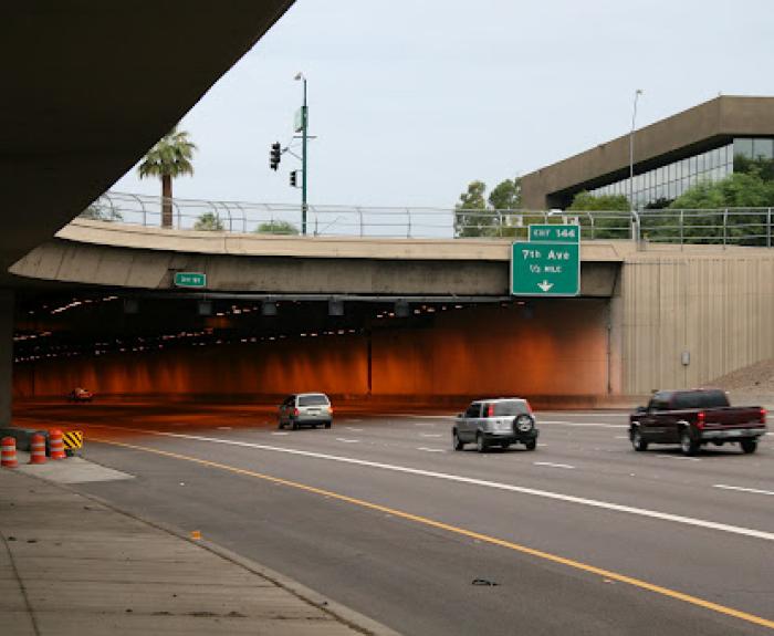 Several vehicles drive toward and into a tunnel under an overpass on a multi-lane road. An overhead sign indicates an exit for 7th Avenue.