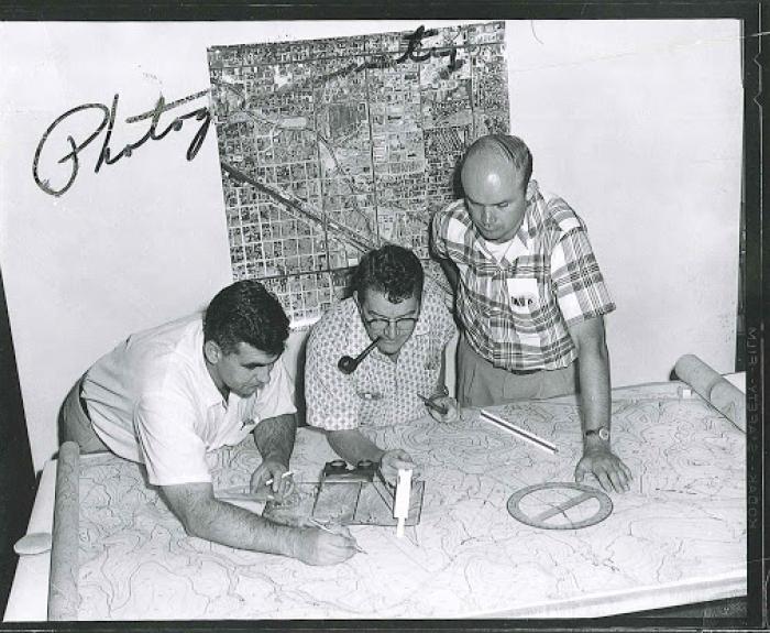 Three men examine a large topographic map on a table, with drafting tools in use; a city map hangs on the wall behind them.