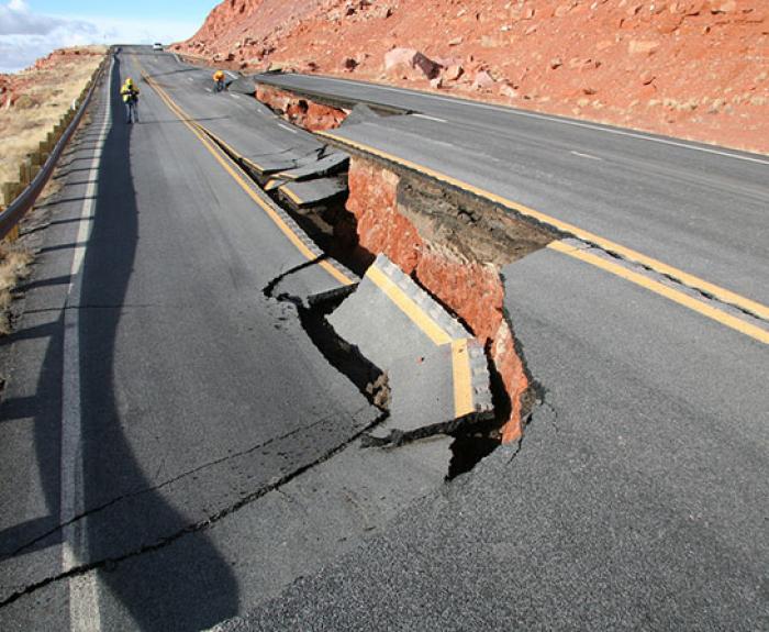 A cracked highway with a large section collapsed, exposing red earth beneath; workers in safety vests are visible in the background near the damaged area.