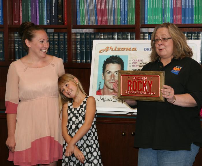 Two women and a young girl stand in front of a large driver license poster and an Arizona license plate.