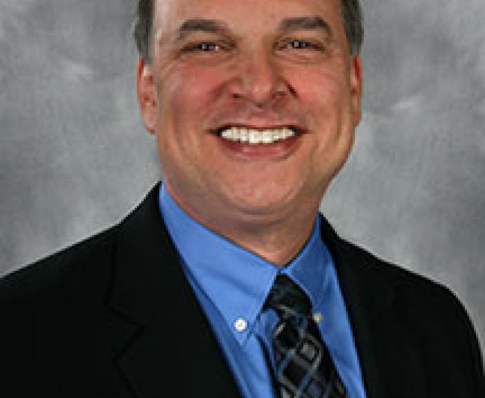 A man in a black suit jacket, blue dress shirt, and patterned tie smiles. He is posed against a gray studio backdrop.