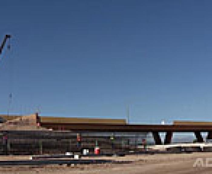 A crane and construction equipment are positioned near an unfinished highway overpass under a clear blue sky.
