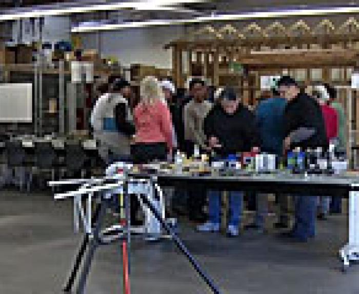 A group of people gathers around a table with various tools and materials in a workshop setting, with shelves and partially built structures in the background.