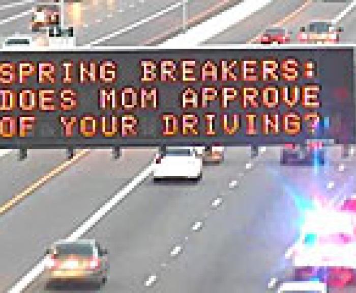 Highway sign displays SPRING BREAKERS! DOES MOM APPROVE OF YOUR DRIVING? with a truck passing by, highlighting a road safety message from Arizona DOT.