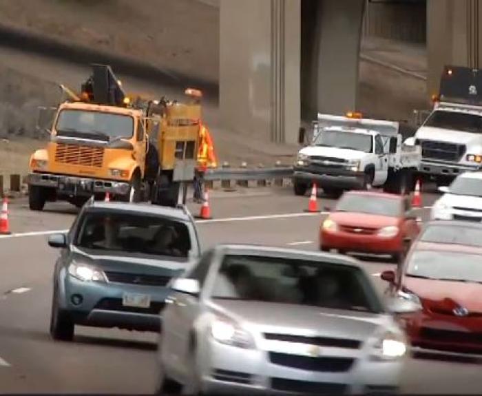 Road construction vehicles and workers on a highway, with traffic cones guiding cars around the work area. Multiple vehicles are moving through the scene.