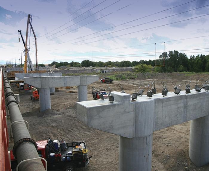 Concrete bridge supports are under construction at a worksite with cranes, vehicles, and equipment; trees and power lines are visible in the background.