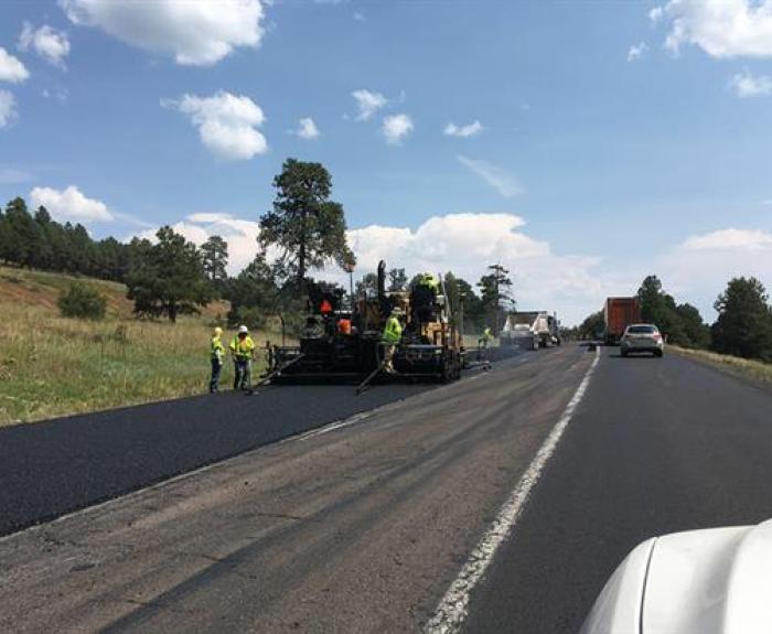 Road construction workers operate paving machinery to lay new asphalt on a highway, with traffic passing by and trees lining the road under a partly cloudy sky.