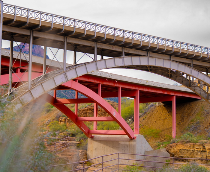 Two steel bridges with arched supports and railings cross over a dry, rocky landscape with sparse vegetation under a cloudy sky.