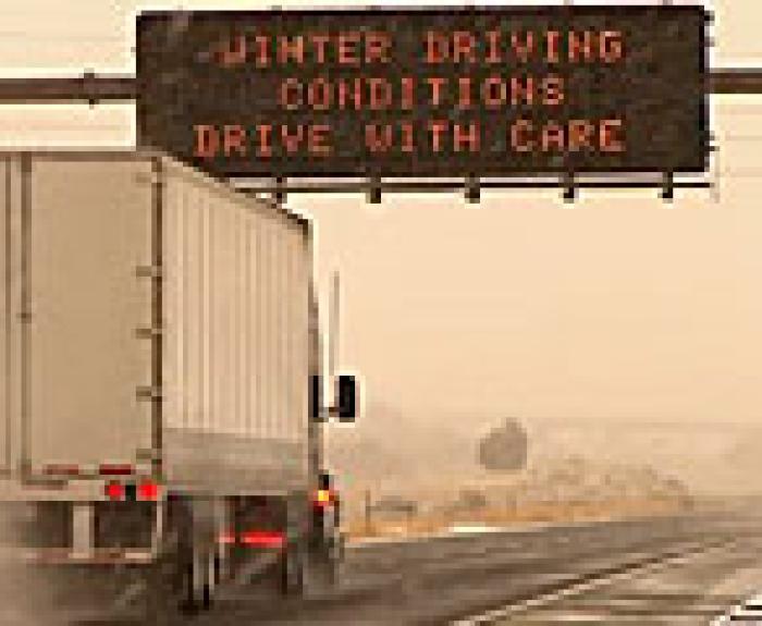 truck driving through a snowy highway with a road sign above saying winter driving conditions drive with care