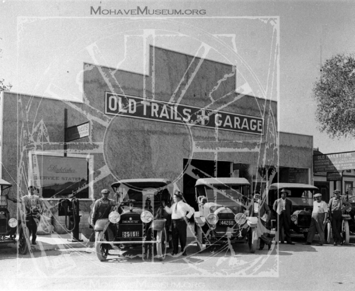 Black and white image of Old Trails Garage. Multiple old vehicles, including Model T's, are parked in front while men stand beside the vehicles.