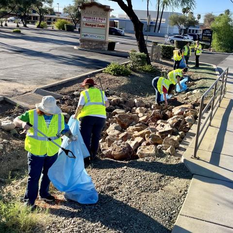 People in yellow reflective safety vests pick up litter on the side of a road and near a parking lot.